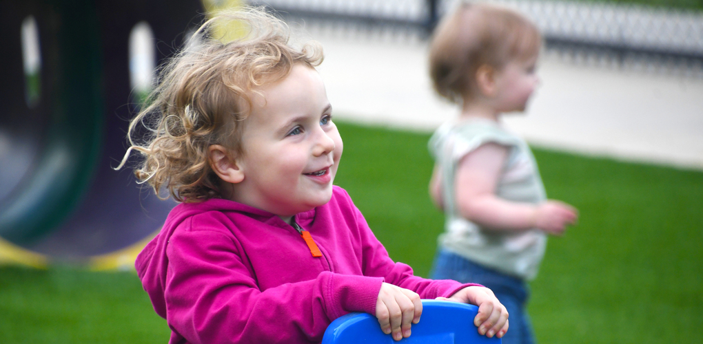 Two children playing on a playground, with one smiling while riding a toy and the other in the background.