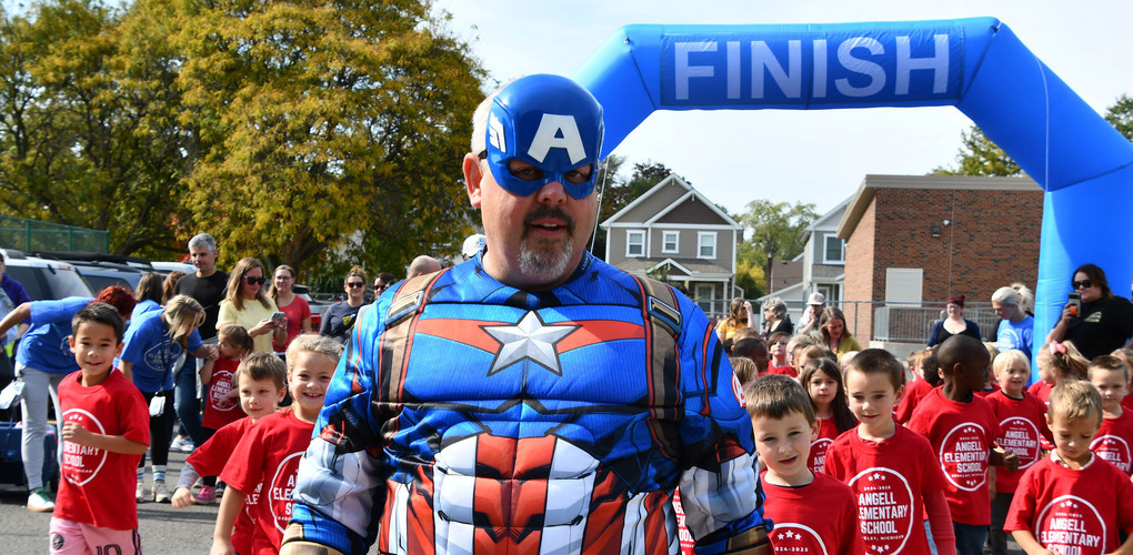 A person in a Captain America costume leads a group of children through a finish line at an event, with spectators watching on both sides