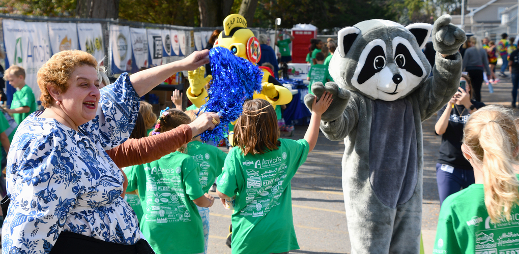 A group of people, including a mascot dressed as a racoon, participating in a lively outdoor community event with someone holding pompoms and others wearing bright green t-shirts.