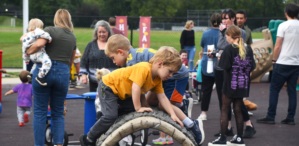 Children and adults at a playground, some kids playing on a tire-based equipment while others interact in the background.