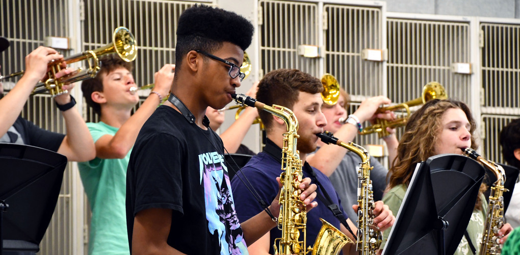 A group of high school students enthusiastically playing saxophones and trumpets in a band rehearsal room.