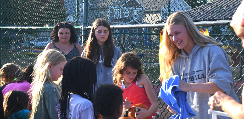 Group of children and adults participating in an outdoor community event, with one person handing out pumpkins to the children.