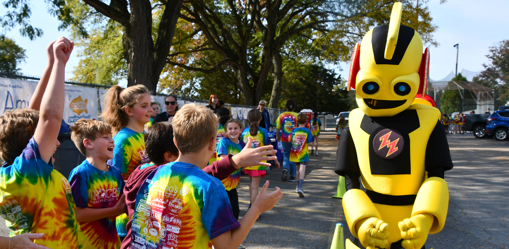 Students cheering on a robot mascot as he dances in a parking lot with teachers spectating in the behind them