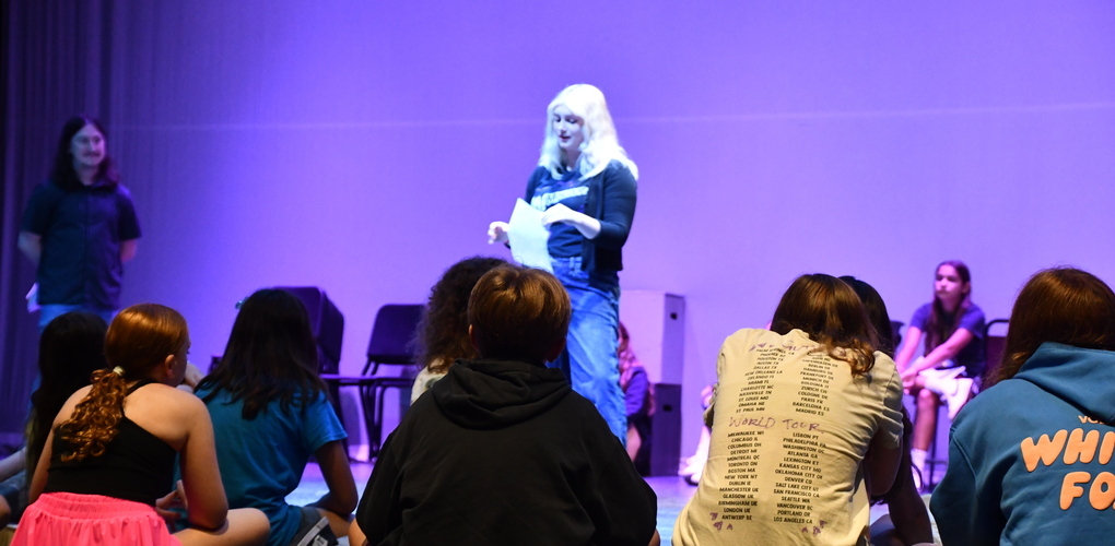 A student giving a performance on stage while other students sit watching with teacher to the left overlooking