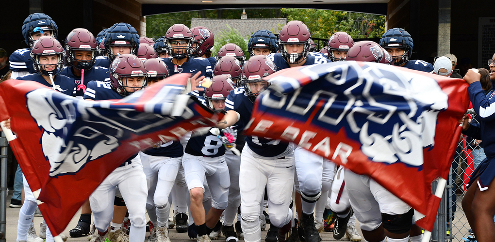 A group of high school football players in maroon and blue uniforms rush out from under a stadium tunnel, enthusiastically waving flags that reads "Bears"