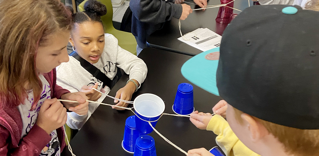 Group of students engaging in a teamwork activity involving stacking cups using strings in a classroom setting.