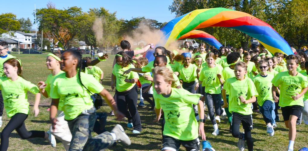 Students happily running in green colored shirts under a rainbow colored arch with colorful smoke above them