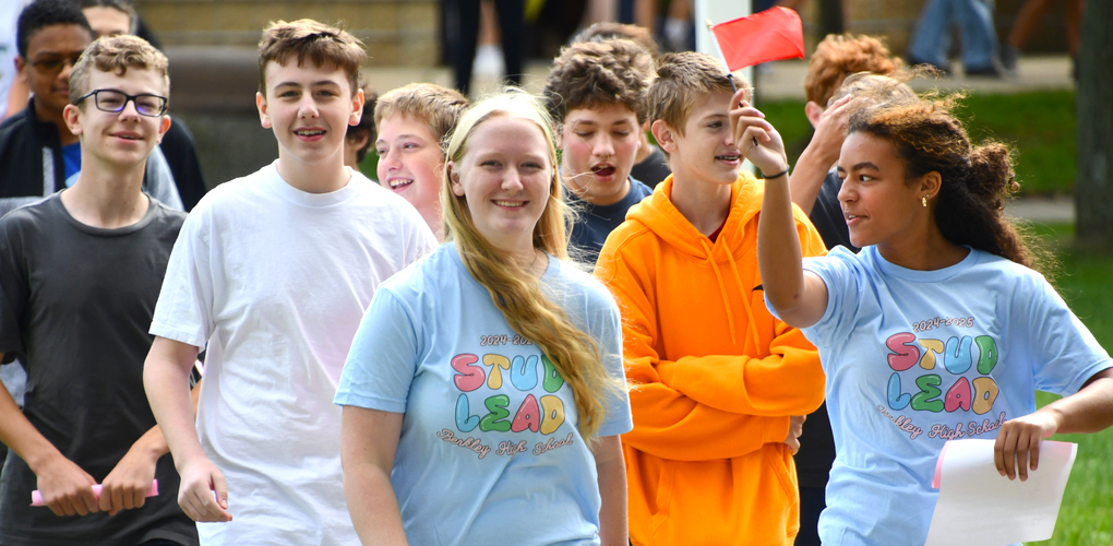 A group of students wearing 'STUD LEAD' t-shirts walking together outdoors, engaged in a lively discussion and smiling.