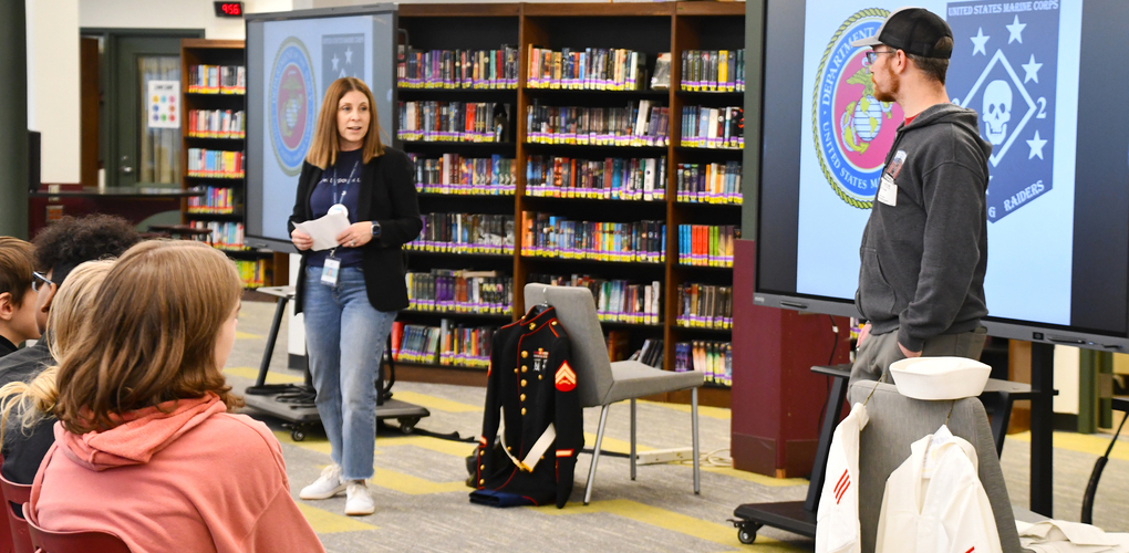 Two people presenting in a library with a focus on U.S. Marine Corps, as seen on the screen behind them, to an audience of students.