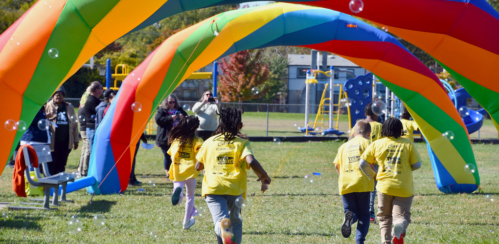 Children in yellow shirts running through a colorful parachute arch with bubbles floating in the air at a sunny playground.