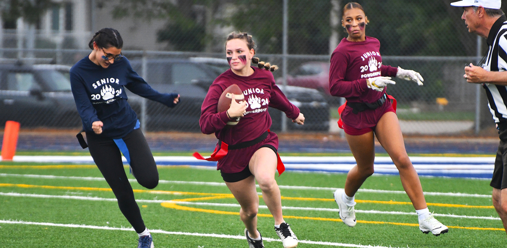 Three high school students are engaged in a flag football game on a sports field, with one student in pursuit of another who is carrying the ball, while a referee watches nearby. The students are wearing sweatshirts labeled "Senior 2024" and "Junior 2024.