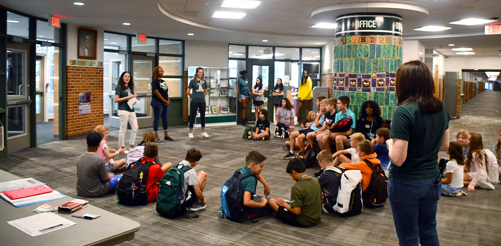 Several students with backpacks sitting on the floor in a school hallway, listening to a teacher standing and speaking. Other teachers and students stand scattered around the spacious area.